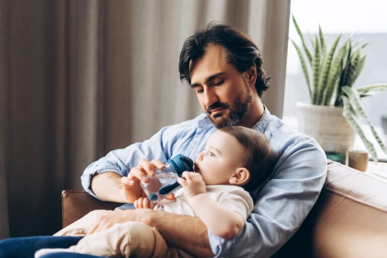 Smiling dad holding infant boy while he drinking water, sitting in living room at home